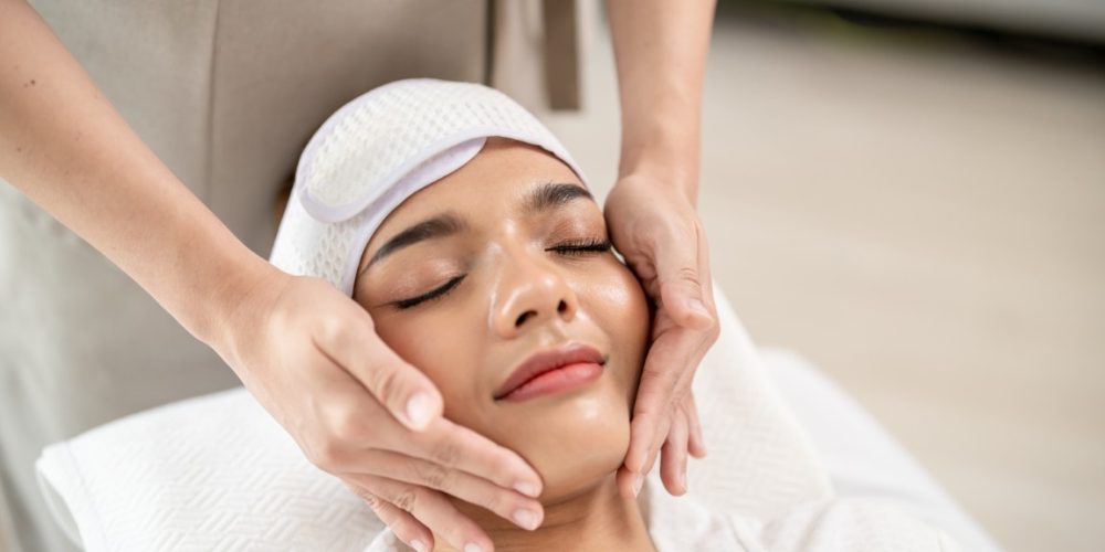 Beautiful woman getting head and facial massage treatment at beauty spa. Attractive girl lying down on massage bed, getting relaxing physiotherapy service from masseuse for body care in spa clinic.